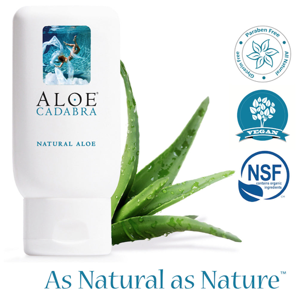 A bottle of Aloe Cadabra, a natural aloe based lubricant with an image of a woman swimming underwater. The packaging is white with blue text. An aloe vera plant is next to the bottle. On the bottom of the image, the brands slogan reads, "As Natural as Nature". To the right are thee labels that describe the product as being all natural, free of glycerin and paraben, vegan, and made with organic ingredients.
