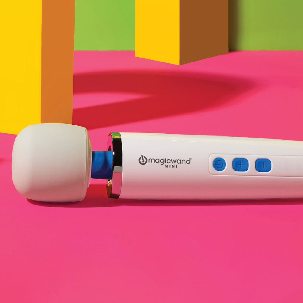 This image shows a close up view of the magic wand mini's head and control buttons on a pink, yellow, and green geometric background. The magic wand mini is white and has three blue operational buttons: a circular power button to turn the vibration on and off, and two rounded square buttons with a plus sign on one and a minus sign on the other to adjust the speed.